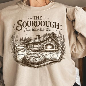 May include: Beige sweatshirt with a graphic of a sourdough bread scene. The design includes a cottage, bread, a jar labeled "Sourter", and the text "THE SOURDOUGH Flour. Water Salt. Time."