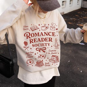 May include: A beige sweatshirt with the text "Romance Reader Society" in burgundy, along with illustrations of books, a rose, and a teacup. The person is wearing a brown baseball cap with "La Paris" printed on it.