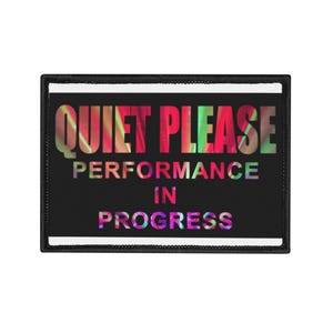 May include: A rectangular black patch with the words "QUIET PLEASE PERFORMANCE IN PROGRESS" in colourful, stylised text. The patch has a black border and a white edge.