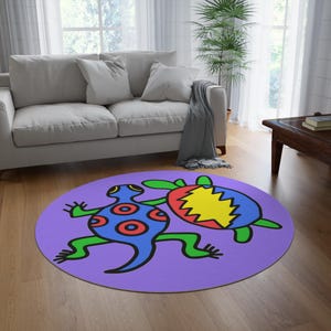 Op de afbeelding: Een ronde, paarse vloerkleed met een grillig ontwerp van twee cartoonachtige wezens. Eén is blauw met rode cirkels, de andere een schildpad met een rood en geel schild. Het kleed ligt op een houten vloer in een woonkamer.