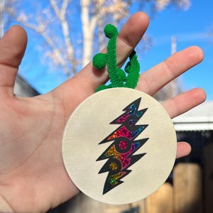 May include: A white round ornament with a rainbow glitter lightning bolt design. The ornament is attached to a green tassel.