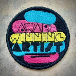 Puede incluir: Parche bordado con las palabras "AWARD WINNING ARTIST" en negro, rosa, amarillo y azul. El parche tiene un borde negro y un diseño colorido en capas. El texto está en negrita, sin serifa.