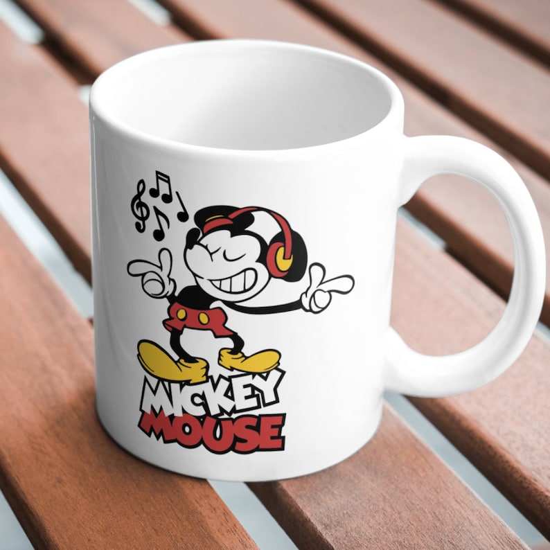 Puede incluir: Taza de cer&aacute;mica blanca con una imagen de dibujos animados en blanco y negro de Mickey Mouse con auriculares y cantando. El texto "MICKEY MOUSE" est&aacute; impreso en rojo y negro debajo de la imagen.
