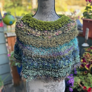 May include: A textured, multi-colored knit poncho featuring a gradient of moss green, teal, deep blue, and purple yarns. The garment has a fuzzy, looped surface with interspersed flecks of pink and metallic threads, draped over a mannequin.