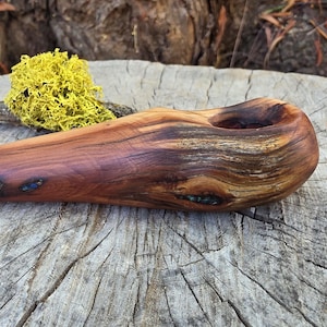 May include: A hand-carved wooden pipe featuring a smooth, polished finish with visible natural wood grain patterns, deep brown and reddish tones, and small inlaid blue and green stone accents embedded within the textured wood surface.