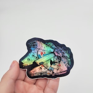 May include: A holographic sticker of the Millennium Falcon spaceship from Star Wars. The sticker is a vibrant rainbow color and has a black background with stars.