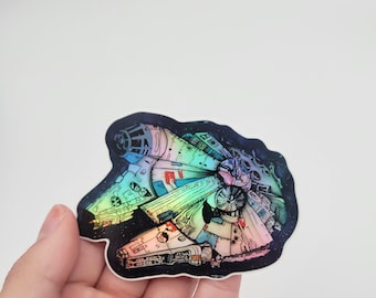 Może przedstawiać: Holograficzna naklejka statku kosmicznego Millennium Falcon z Gwiezdnych Wojen. Naklejka jest w żywych kolorach tęczy i ma czarne tło z gwiazdami.