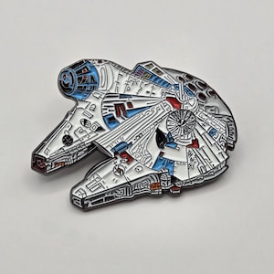 May include: A white, blue, and red enamel pin of the Millennium Falcon spaceship from Star Wars. The pin is detailed with black outlines and has a shiny finish.