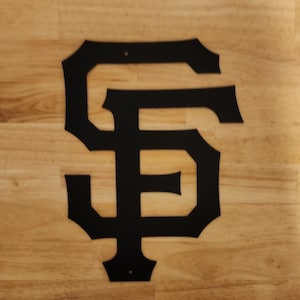 Puede incluir: Corte de metal negro del logotipo de los Gigantes de San Francisco, "SF", en una fuente estilizada. El logotipo está sobre una superficie de madera marrón claro.