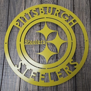 May include: A yellow metal sign featuring the Pittsburgh Steelers logo. The sign is circular with the words "PITTSBURGH" and "STEELERS" cut out around the logo. The logo includes the team name and three yellow hypocycloids.