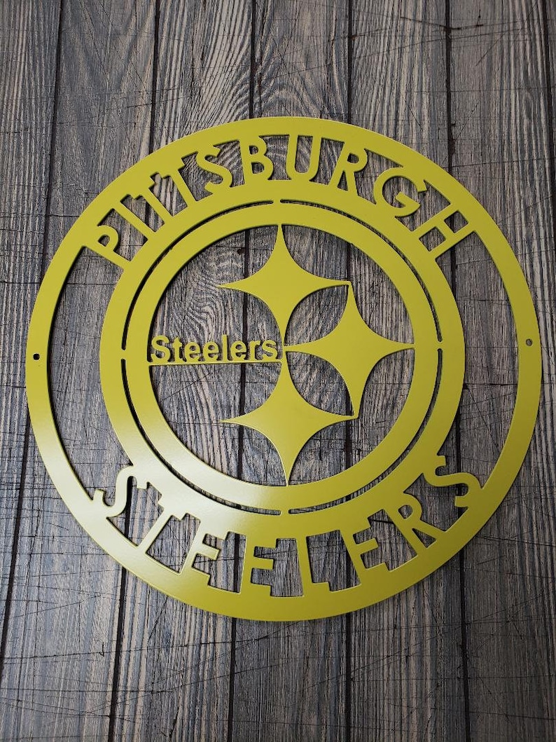 May include: A yellow metal sign featuring the Pittsburgh Steelers logo. The sign is circular with the words "PITTSBURGH" and "STEELERS" cut out around the logo. The logo includes the team name and three yellow hypocycloids.