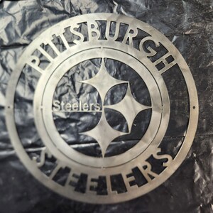 May include: A silver metal sign featuring the Pittsburgh Steelers logo. The team name and city name are cut out around the logo's perimeter. The logo includes the team name and a four-pointed star design.