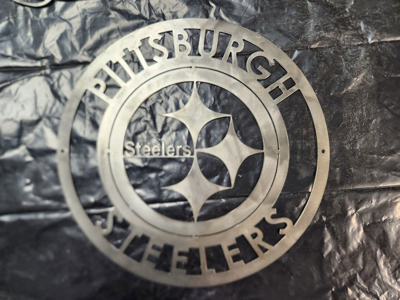 May include: A silver metal sign featuring the Pittsburgh Steelers logo. The team name and city name are cut out around the logo's perimeter. The logo includes the team name and a four-pointed star design.