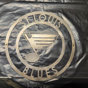 May include: Metal wall art featuring the St. Louis Blues logo. The logo is a stylized note with wings and the words "St. Louis Blues" around the outside.