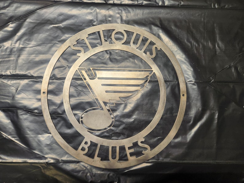 May include: Metal wall art featuring the St. Louis Blues logo. The logo is a stylized note with wings and the words "St. Louis Blues" around the outside.