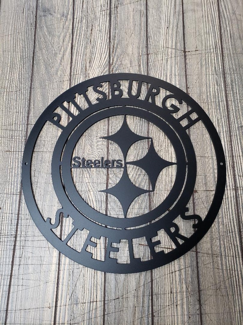 May include: Black metal Pittsburgh Steelers sign. The sign features the team's logo, a circular design with the team name and the word "Steelers" in the center. The sign is designed to be mounted on a wall.