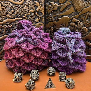 May include: Two crocheted drawstring pouches in variegated shades of purple, magenta, and blue, featuring a textured dragon scale pattern. Arranged in front are seven metallic gold-toned polyhedral dice with intricate black engraved numbering.