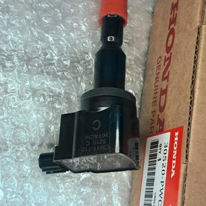 Puede incluir: Una bobina de encendido negra con una tapa protectora roja en la punta, con el texto CM11-110 5215 C HITACHI impreso en el lateral. Está colocada junto a una caja de cartón marrón etiquetada HONDA GENUINE PARTS con un código de barras y el texto 30520-PWC-S01.