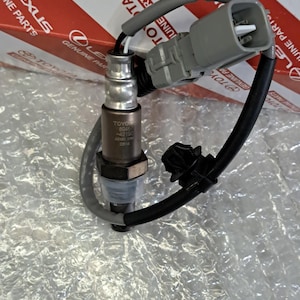 Può includere: Un sensore di ossigeno metallico con un connettore elettrico in plastica grigia e cablaggio nero. Il corpo cilindrico presenta il testo TOYOTA 89467-42190 DENSO 1700 03514. Il componente include una clip in plastica nera sul cablaggio.