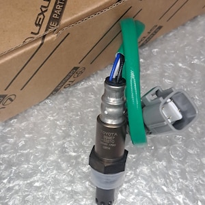 May include: A metallic automotive oxygen sensor featuring a dark grey cylindrical body with the text TOYOTA 89467-33210 DENSO 1780 03R14 printed on it. The sensor includes a green protective wire sleeve, blue and black internal wires, and a grey connector.