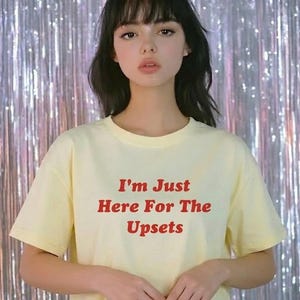 Puede incluir: Camiseta amarillo claro con el texto rojo "I'm Just Here For The Upsets". La persona lleva la camiseta. El fondo es una cortina plateada brillante que crea un efecto reflectante.