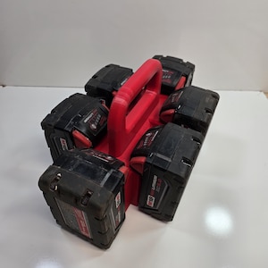 May include: A red battery holder with a handle, holding six black M18 Lithium XC 5.0 batteries. The batteries are arranged around the holder, with the red handle at the top. The batteries are for power tools.