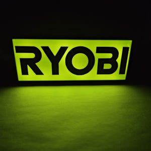 May include: Illuminated sign with the word "RYOBI" in bold, black letters against a bright green background. The sign is rectangular and glows in the dark, with a dark background.
