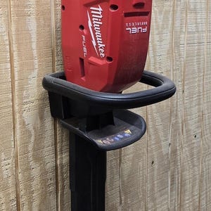 Puede incluir: Una podadora de setos inalámbrica roja y negra con un mango y protector de cuchilla negros. La podadora está montada en una pared de madera. La marca "Milwaukee" es visible en la herramienta.