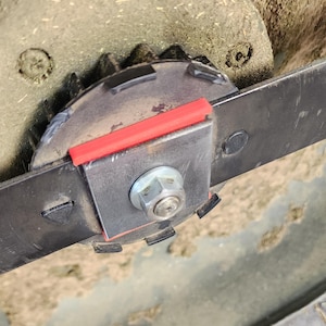 Pode incluir: Close-up de um conjunto de lâmina de cortador de grama. A lâmina é fixada a uma placa de metal com um espaçador vermelho. Um parafuso grande prende a lâmina. O conjunto é cercado por grama cortada.