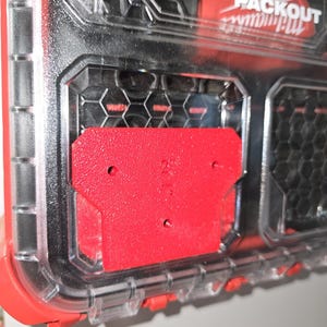 Può includere: Un contenitore di stoccaggio Milwaukee Packout rosso e trasparente. Il contenitore ha una base rossa e un coperchio trasparente, con il logo Milwaukee e il marchio Packout visibili. C'è un componente rettangolare rosso con due fori.