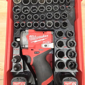 May include: A red Milwaukee FUEL impact wrench kit with a variety of black sockets and two batteries. The kit is housed in a red plastic case with a clear lid. The Milwaukee logo is visible on the wrench and batteries.