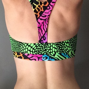 Puede incluir: Un sujetador deportivo de color verde ne&oacute;n, rosa, naranja y negro con estilo de espalda de nadador. El sujetador tiene un patr&oacute;n geom&eacute;trico con detalles de estampado animal.