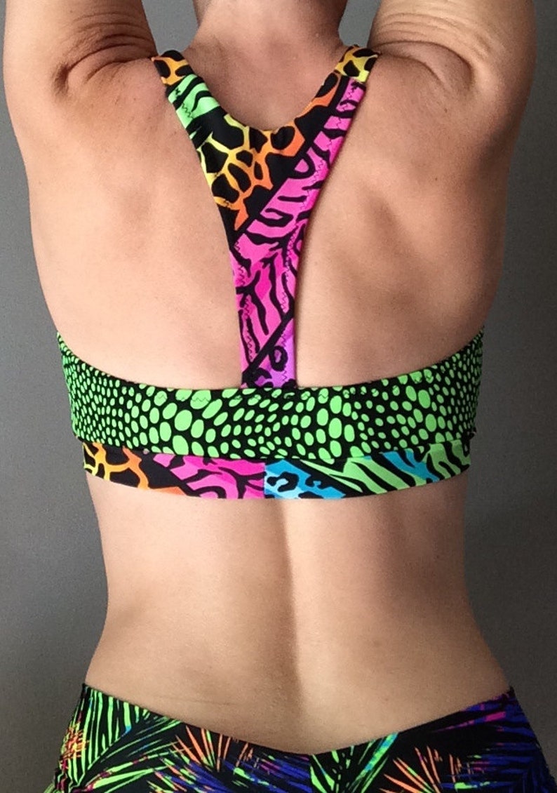 Puede incluir: Un sujetador deportivo de color verde ne&oacute;n, rosa, naranja y negro con estilo de espalda de nadador. El sujetador tiene un patr&oacute;n geom&eacute;trico con detalles de estampado animal.
