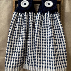 May include: Two navy blue and white checkered kitchen towels with a crocheted top and large white button closure. The towels are hanging on a wooden ladder. The bottom of the towels have a white and blue striped pattern.