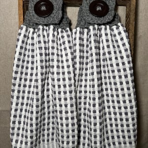 May include: Two hanging kitchen towels with a gray and white checkered pattern. Each towel has a gray crocheted top with a large brown button. The towels are hanging on a wooden ladder.