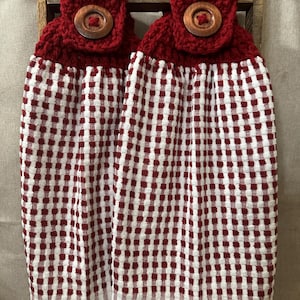 May include: Two red and white checkered kitchen towels with a crocheted top and wooden button closure. The towels are hanging on a wooden rack. The towels have a red and white checkered pattern.