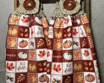 May include: Two hanging kitchen towels featuring a patchwork pattern of pumpkins, autumn leaves, acorns, and the text "HELLO fall" and "pumpkin" in shades of orange, brown, and white. Each towel has a beige crocheted top with a large brown button.