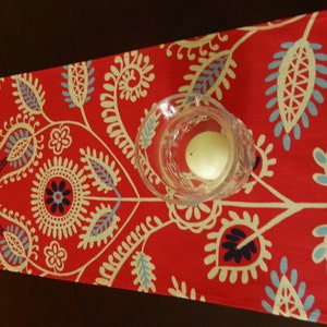 May include: Red table runner with a white and blue floral pattern. A clear glass candle holder sits on the runner.