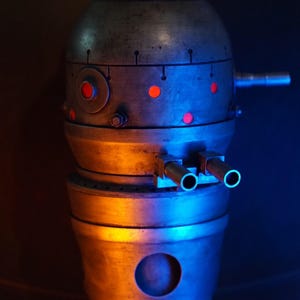 May include: A silver and gray metal robot with red lights and two silver barrels. The robot is lit with blue and orange lights.
