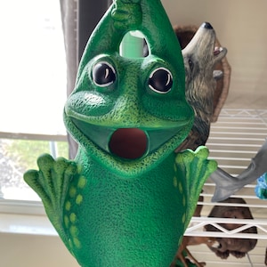 May include: A green frog figurine with a wide open mouth, hanging from a white rope. The frog has large black eyes and a textured surface.