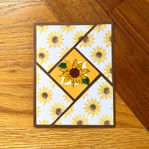 May include: A handmade greeting card with a sunflower theme. The card features a brown border, a white background with yellow sunflowers, and a central yellow square with a detailed sunflower illustration. The card is designed with a geometric pattern.