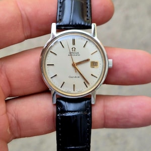Op de afbeelding: Een vintage Omega Genève automatisch horloge met een zilverkleurige kast en een zwarte leren band. De wijzerplaat is crèmekleurig met zwarte markeringen en wijzers. Het horloge heeft een datumvenster op 3 uur.