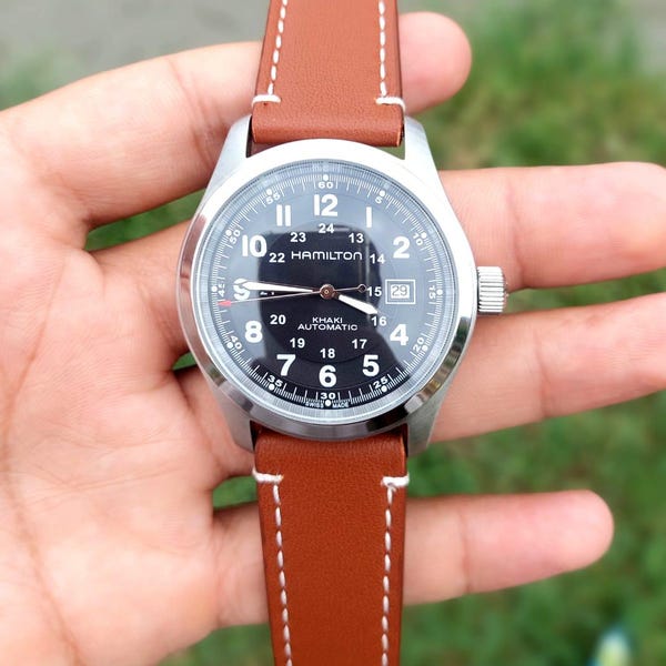 May include: A silver-cased wristwatch with a black face and brown leather strap. The watch face displays the brand name "HAMILTON" and "KHAKI AUTOMATIC". The brown strap has white stitching.