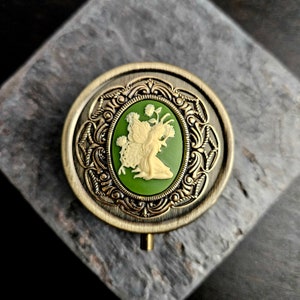 May include: A round, brass trinket box with an ornate design. The lid features a cameo of a fairy with wings, surrounded by green and white flowers.