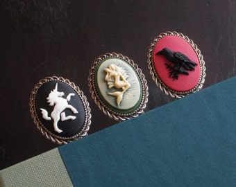 May include: Three silver bookmark holders with black, red, and white cameo designs. The designs are a unicorn, a mermaid, and a crow.