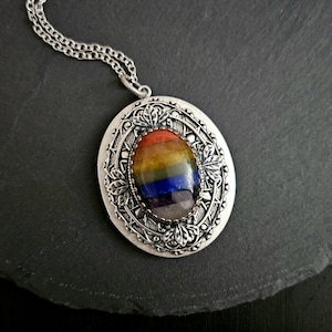 May include: An oval locket pendant on a silver-toned chain. The locket features an intricate, embossed metal frame with leaf-like details surrounding a central, smooth, multi-colored stone inlay with horizontal stripes of red, orange, yellow, green, blue, and purple.