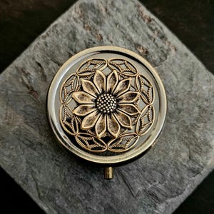 May include: A round, antique-style compact mirror with a gold-colored floral design. The mirror has a dark background and a small handle. The floral design is in the center of the mirror and is surrounded by a decorative border.