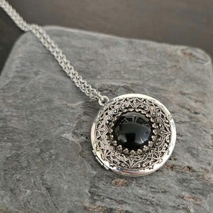 May include: Silver-toned locket necklace with a round, black gemstone center. The locket features an ornate, filigree design around the gemstone and a delicate chain. The necklace is displayed on a gray, textured surface.