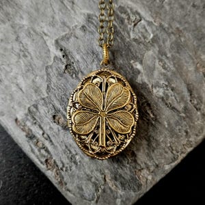 May include: A gold-colored oval pendant necklace with a four-leaf clover design. The pendant is intricately detailed with a filigree border and hangs from a matching chain. The necklace is displayed on a gray, textured surface.