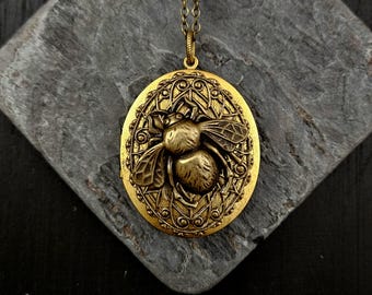 May include: An oval-shaped brass locket pendant featuring a raised bee relief at the center, surrounded by an intricate filigree border. The locket is suspended from a matching brass chain, showcasing a metallic gold finish throughout.